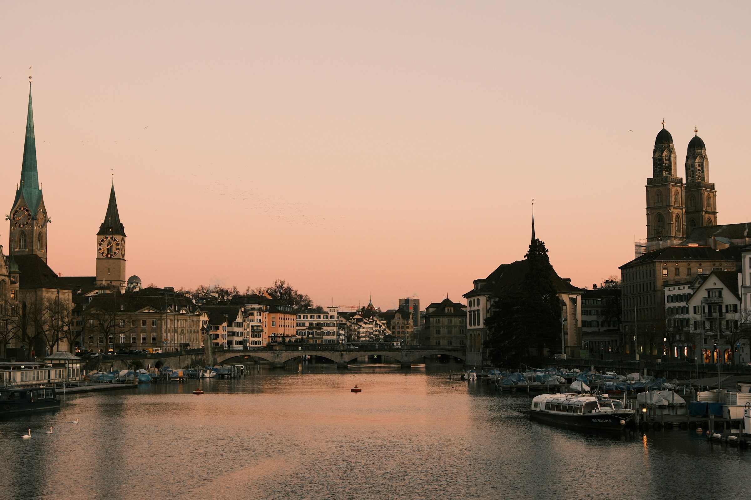 Zurich: the Limmat River, Münsterbrücke, and Fraumünster spire at sunset with reflections.